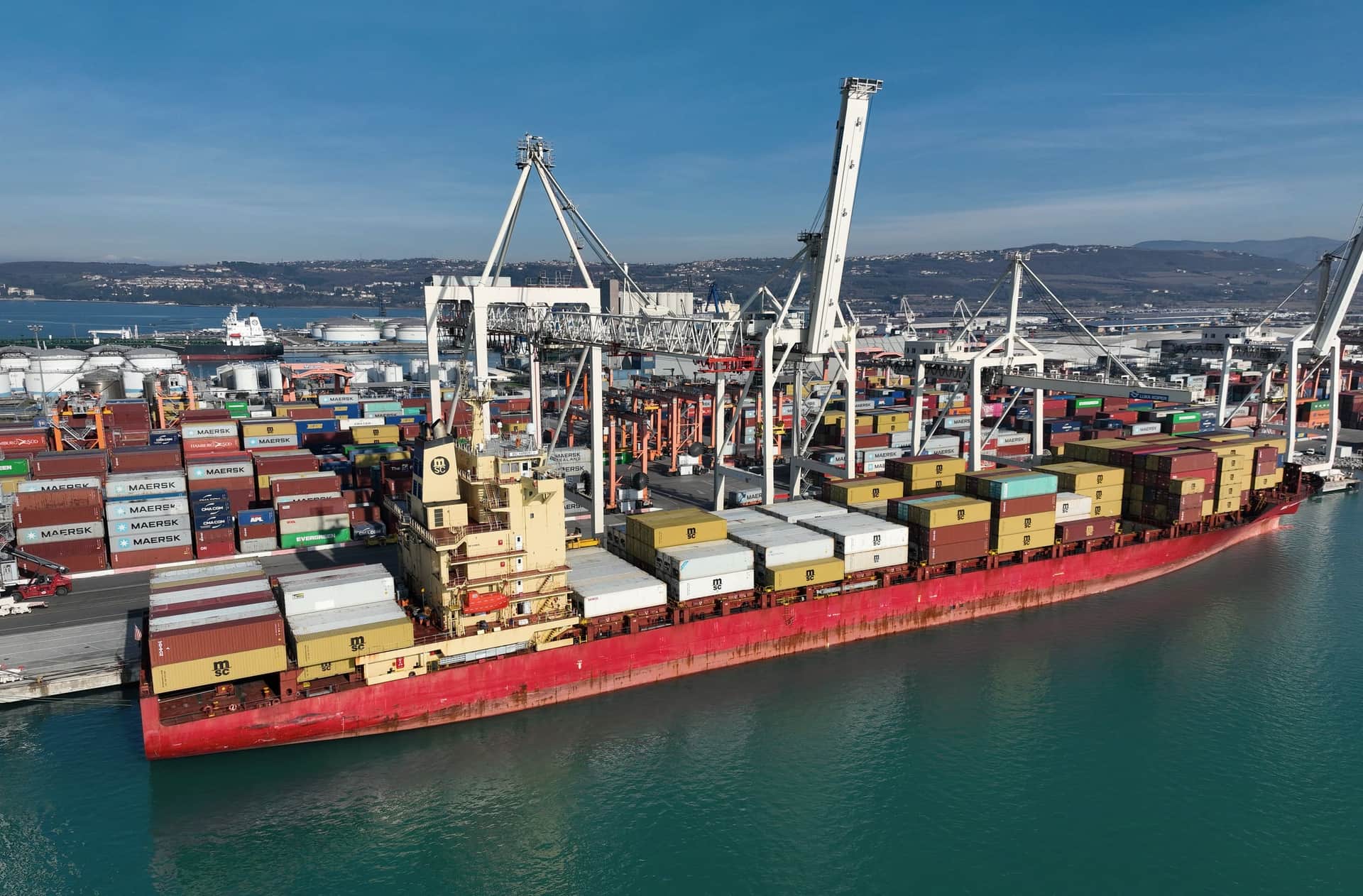 A container ship berthed under quay cranes at the Port of Koper, with straddle carriers on the quay